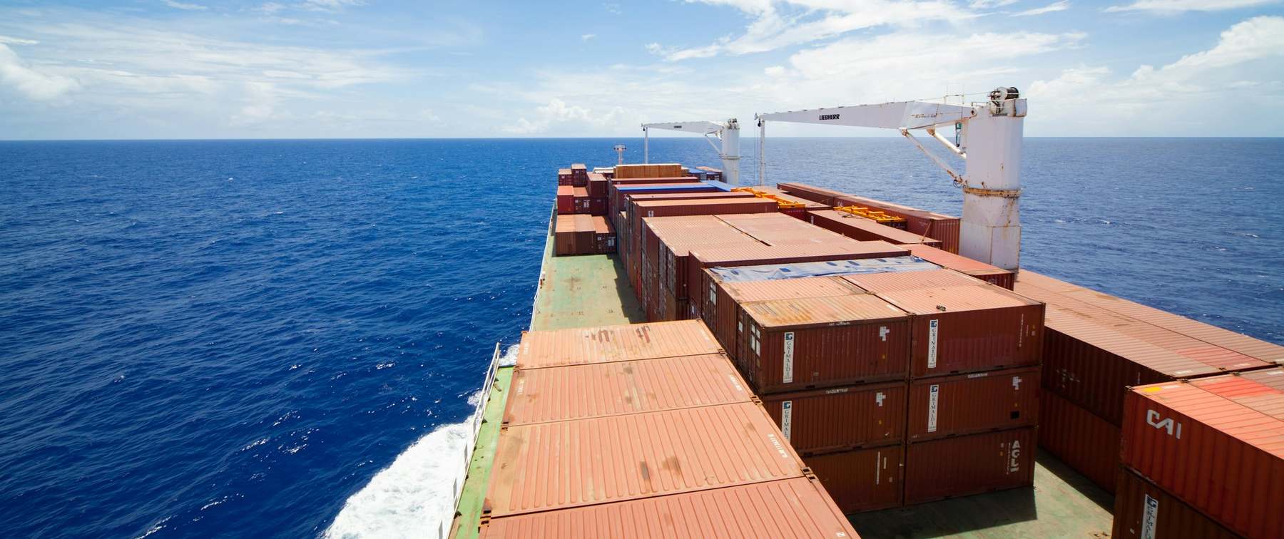 A container vessel under way in open sea, seen looking forward along the cargo deck towards the horizon.